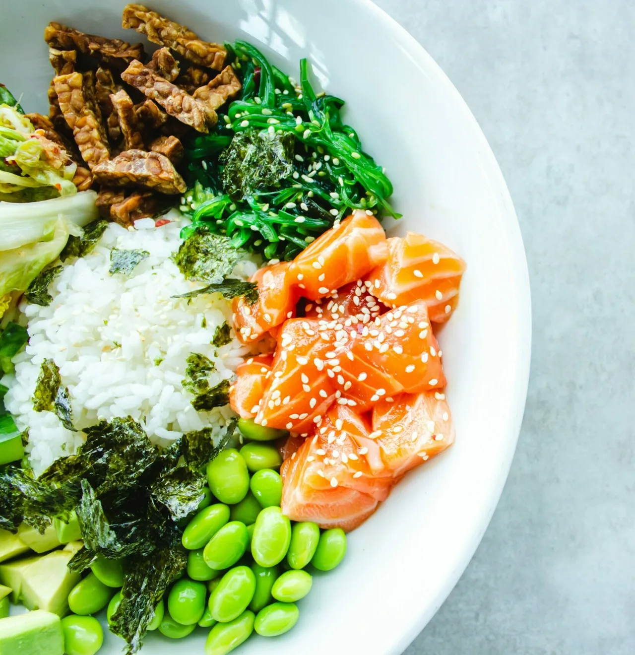 Assiette bowl avec riz blanc, saumon cru tranché parsemé de graines de sésame, légumes verts (algues, edamame, germes), cacahuètes et nori sur fond blanc.