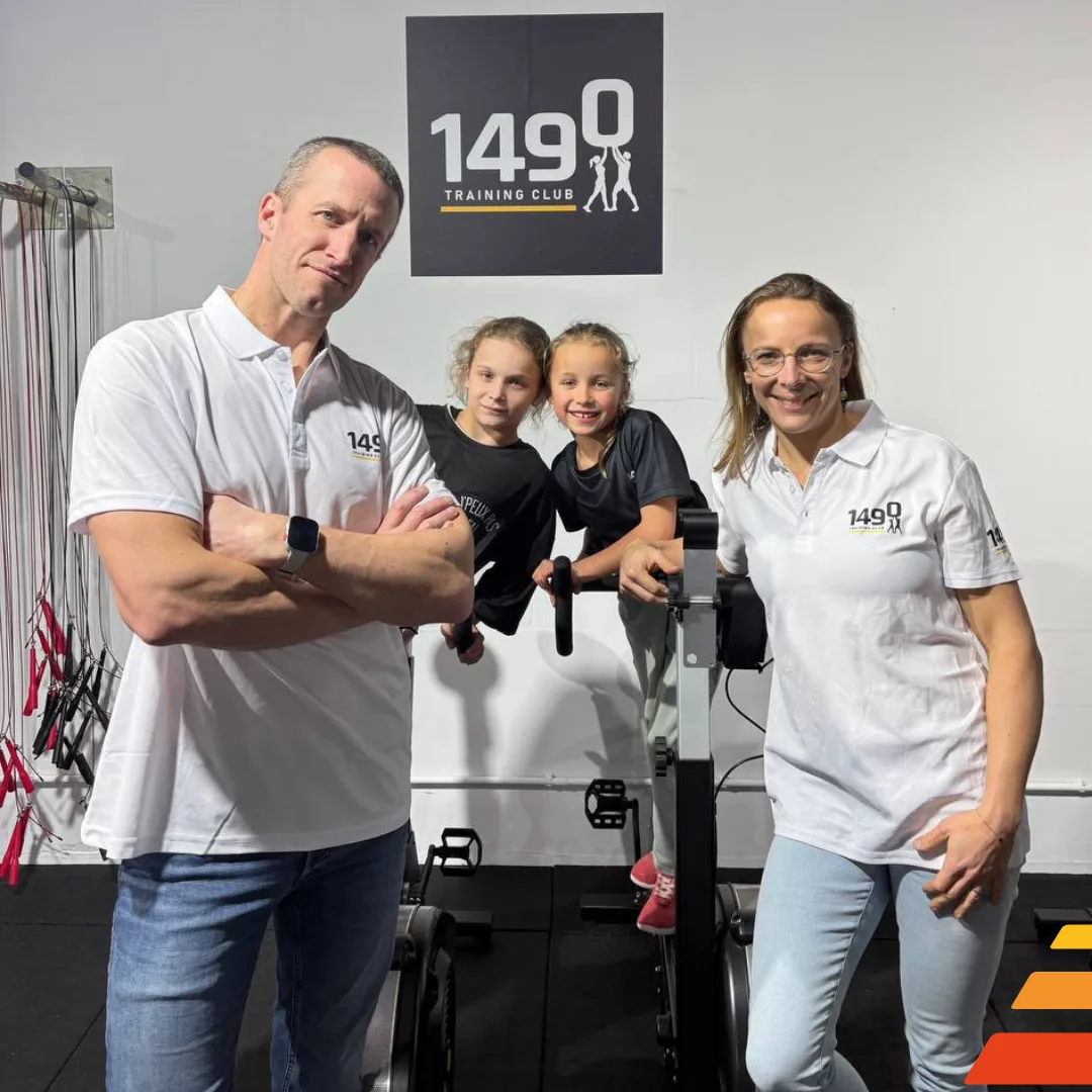 Homme adulte portant un polo blanc au logo du 1490 Training Club, debout face à la caméra dans une salle de sport avec logo du club en arrière-plan sur mur gris.